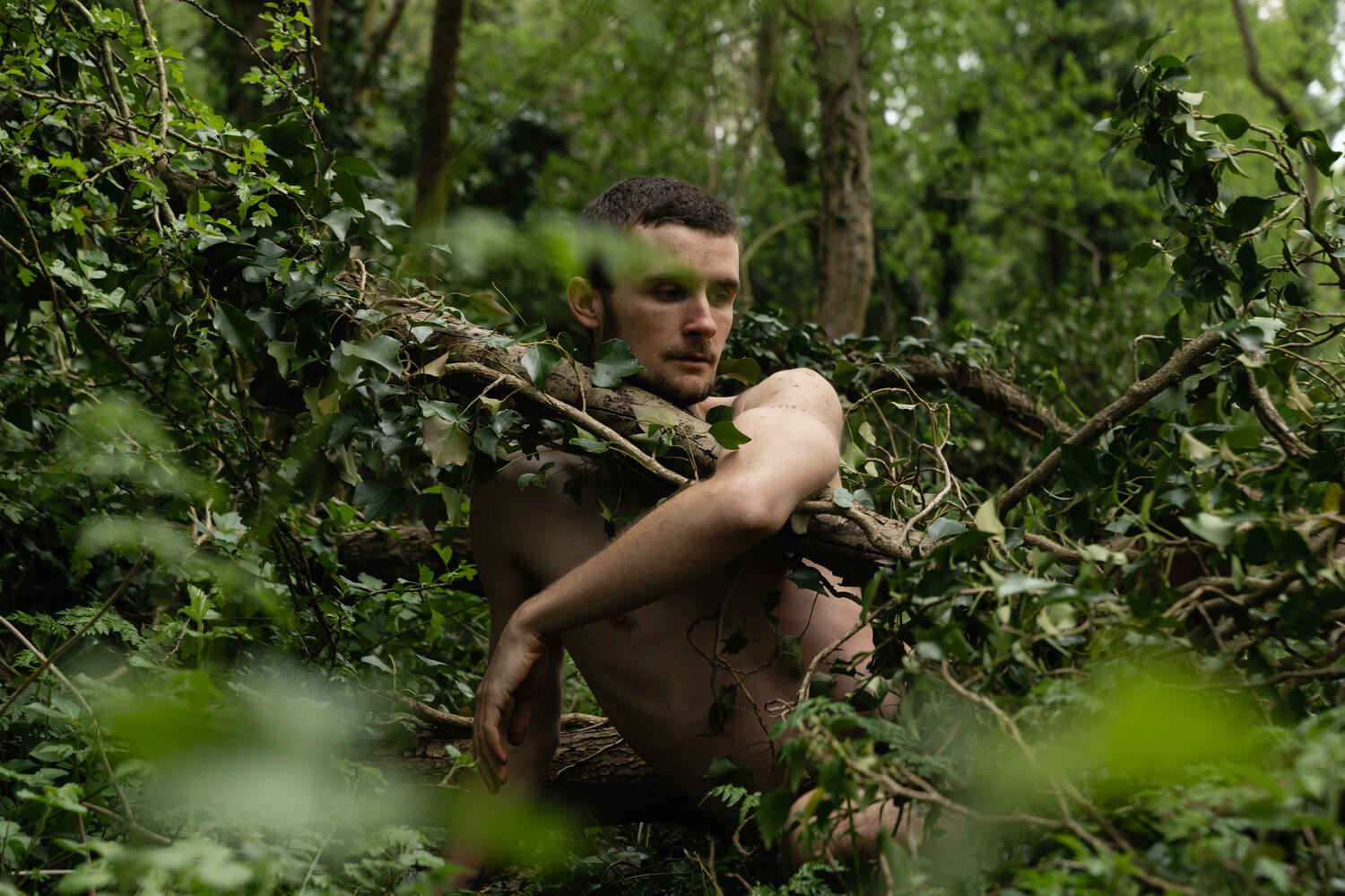 Shaun Hill Embodied Woodland Poet Shaun embracing a tree entwined with ivy with his bicep and bare torso on a forest floor.